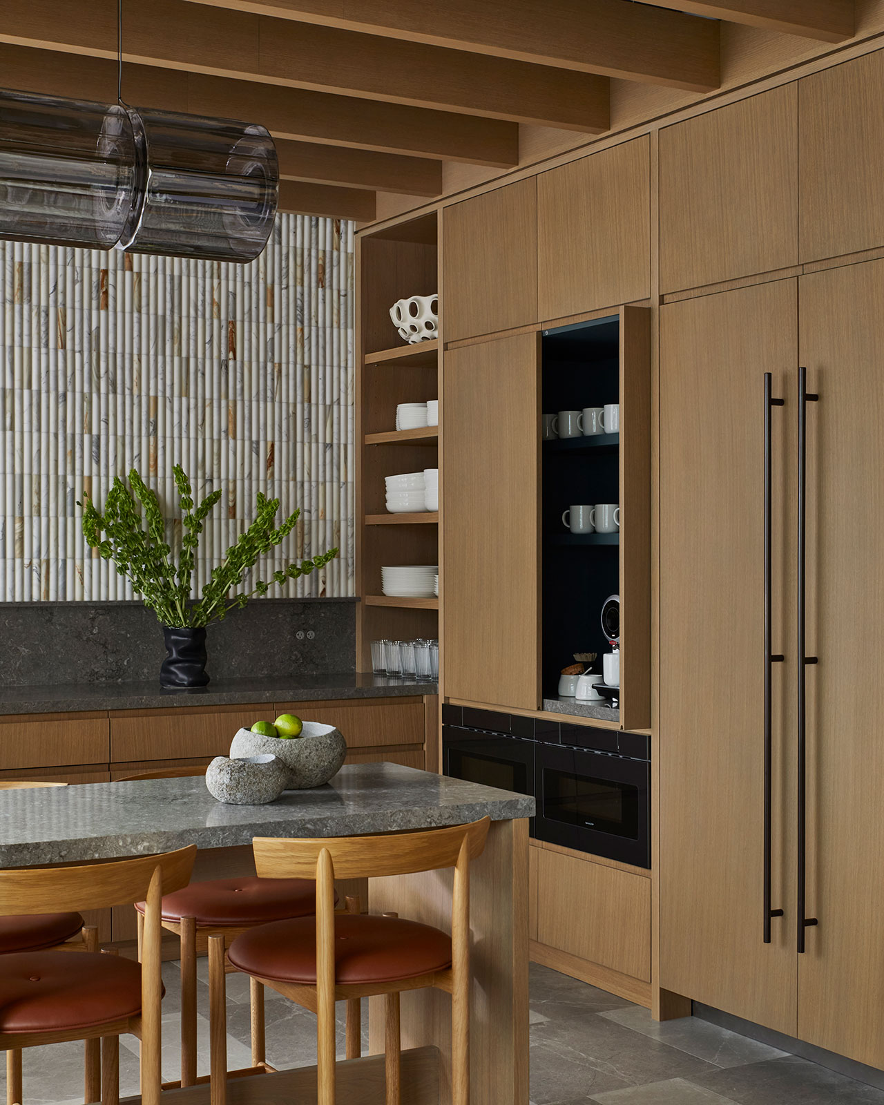 Modern kitchen with wood cabinetry, stone countertops, open shelves with dishes, a built-in appliance, and a vase with green branches on the island. Four wooden chairs surround the island.