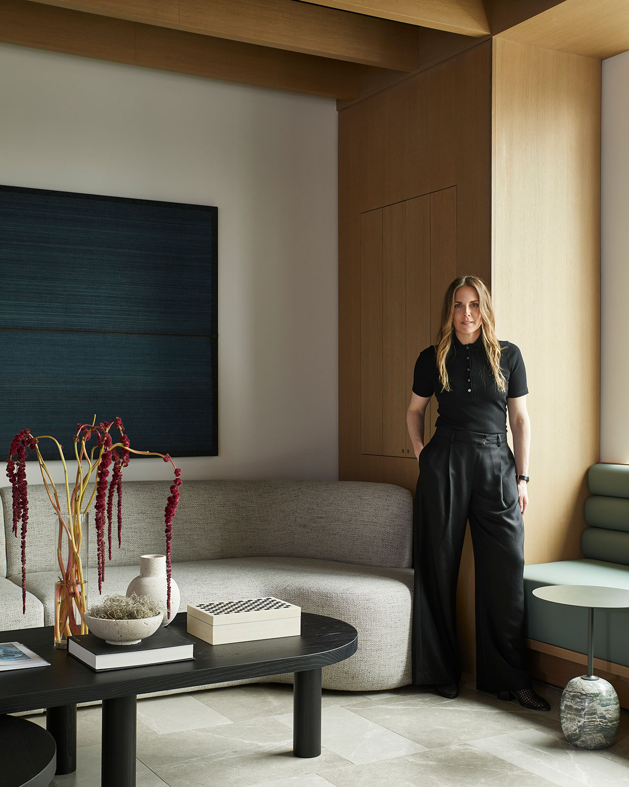 A woman stands in a modern living room with wooden accents, a curved sofa, a round coffee table with vases, and abstract artwork on the wall.