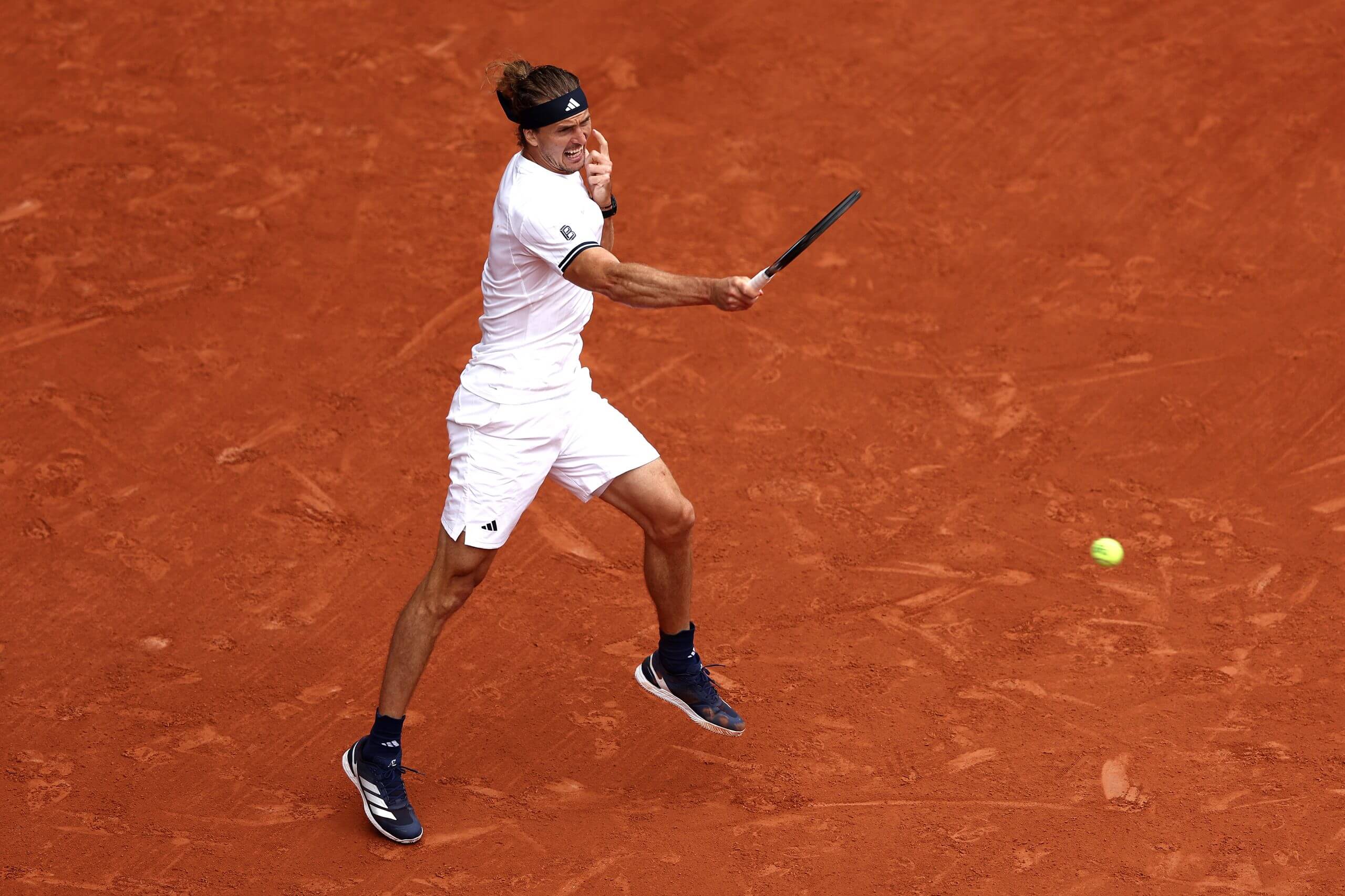 Alexander Zverev hits a forehand on a clay court while wearing a white tennis outfit.
