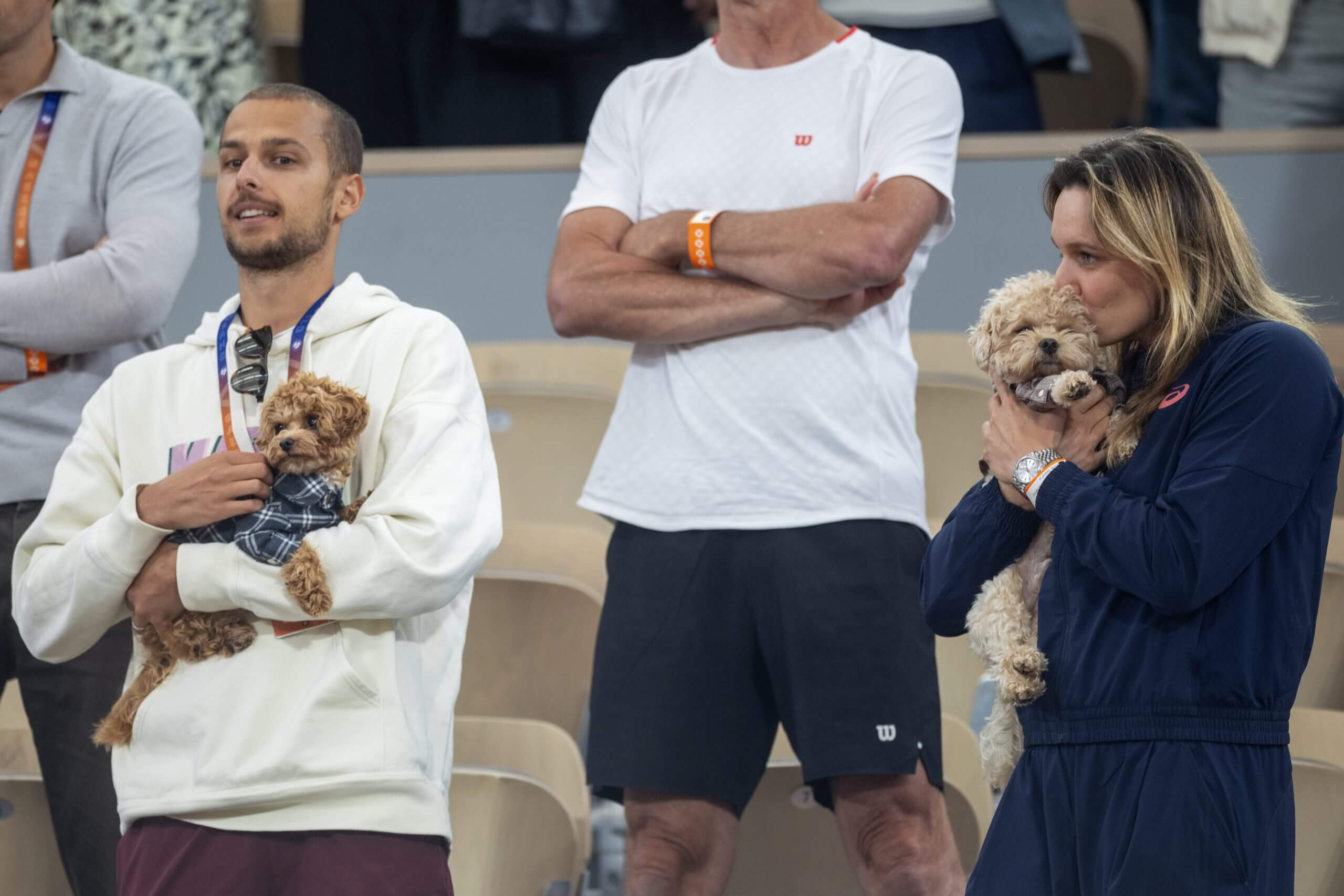 A man holds a small, brown dog while to his right a woman holds a larger, slightly lighter dog, while both are standing in a tennis stadium.