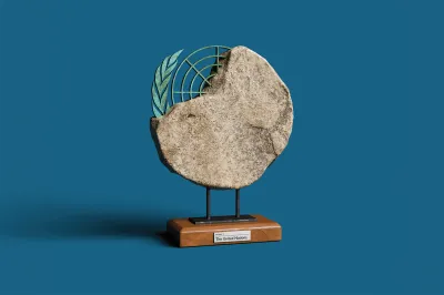 A studio shot of an artifact displayed on a small wooden base against a solid blue background. The artifact features a large, rough-textured stone disk with an uneven edge. Embedded or emerging from the upper-left edge of the stone is a green, weathered metal emblem resembling the United Nations logo, including an olive branch and a partial grid pattern. The stone structure is mounted on two short black metal pillars attached to a polished wooden stand, which has a small white label on the front that reads "EXHIBIT 1: The United Nations".