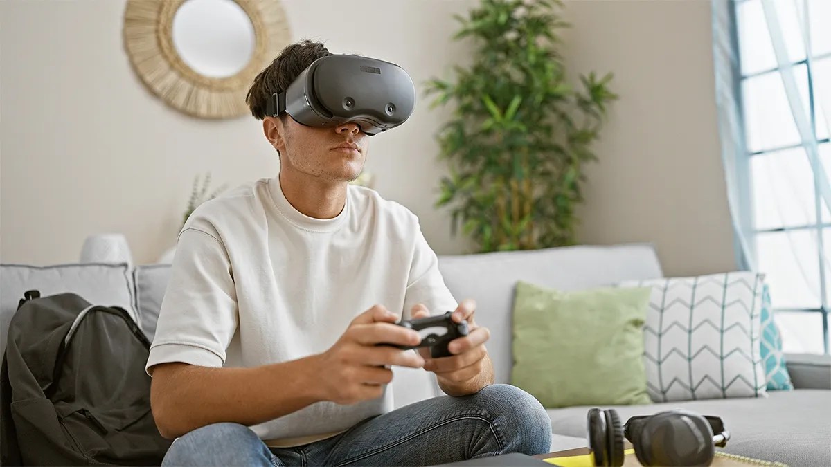 An individual sitting on a couch uses a virtual reality headset while holding a game controller, with a backpack and headphones nearby in a living room setting.