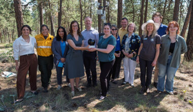 A group of 11 people stand together in a forested area, with the centre two individuals holding a circular, black camera.