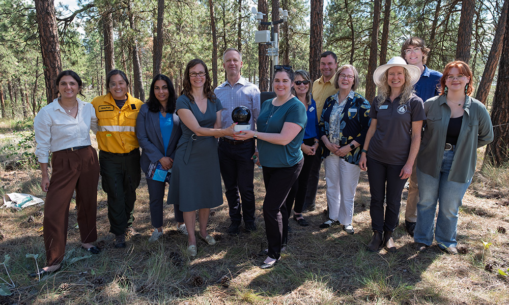 A group of 11 people stand together in a forested area, with the centre two individuals holding a circular, black camera.