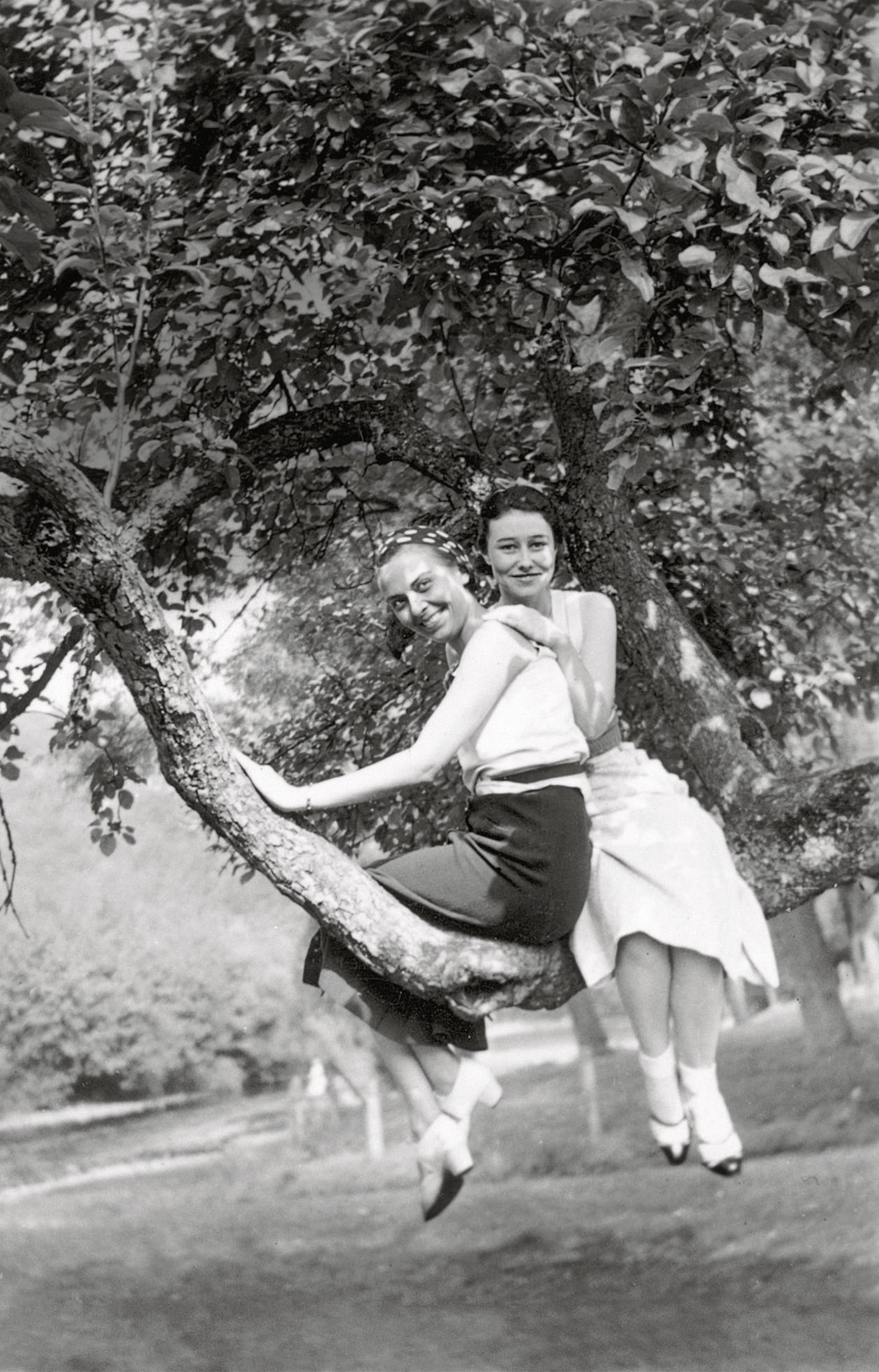 Two women in vintage clothing sit side by side on a curved tree branch, smiling at the camera. Lush foliage surrounds them, and sunlight filters through the leaves, creating a serene, joyful outdoor scene.