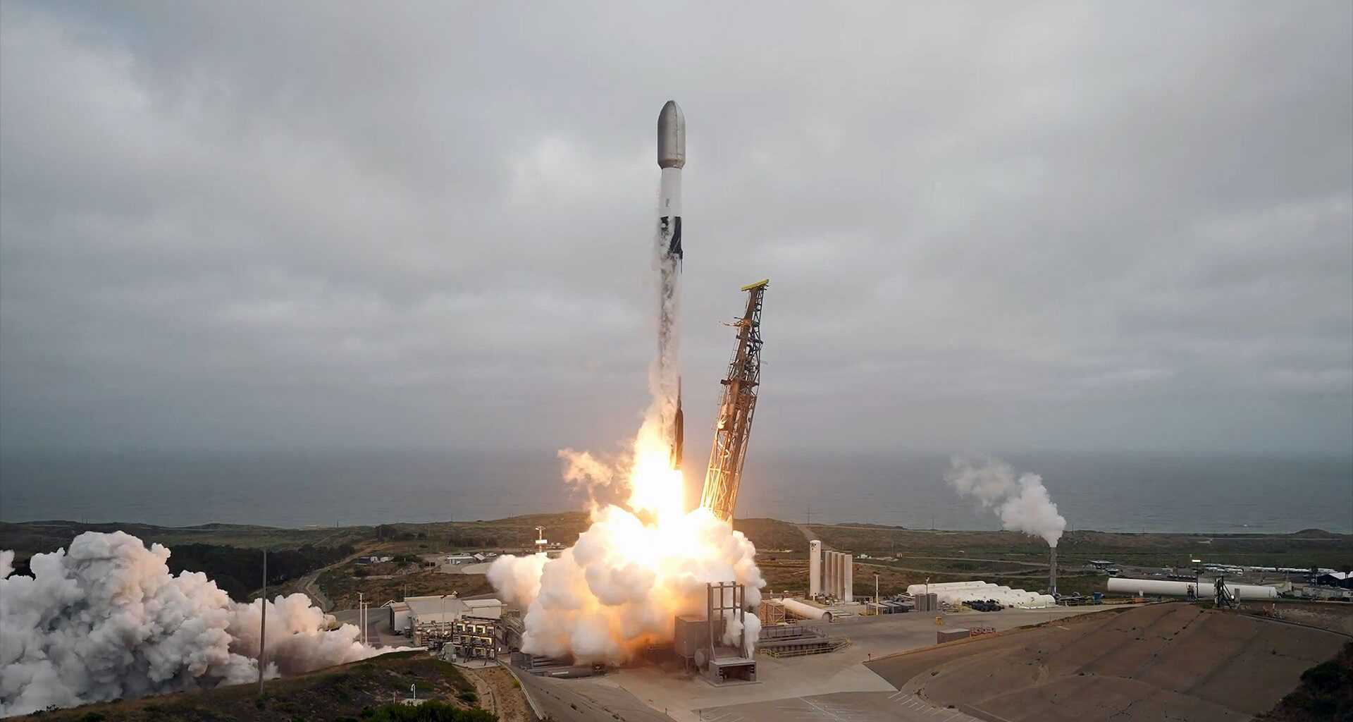 a white and black rocket lifts from a sea-front launch pad into an overcast sky