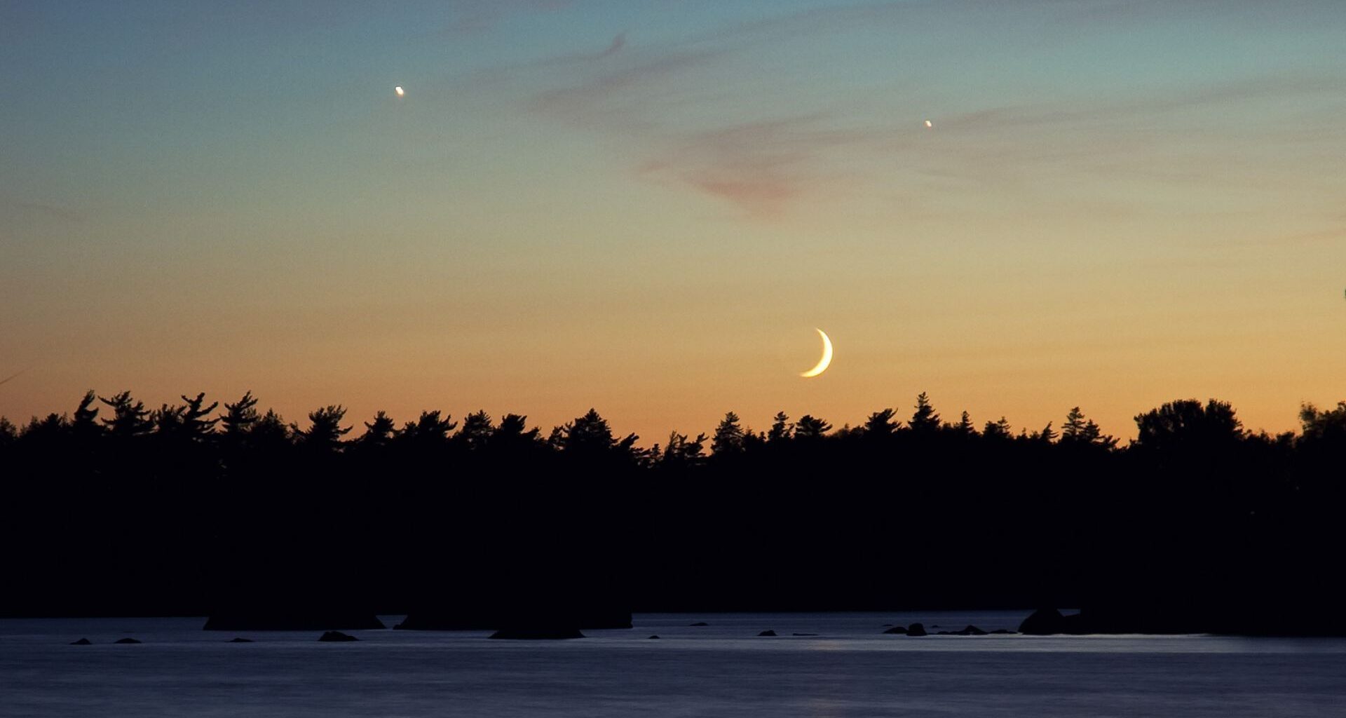 A crescent moon shines in the evening sky above a forest as two planets glow overhead in the peach colored sky.