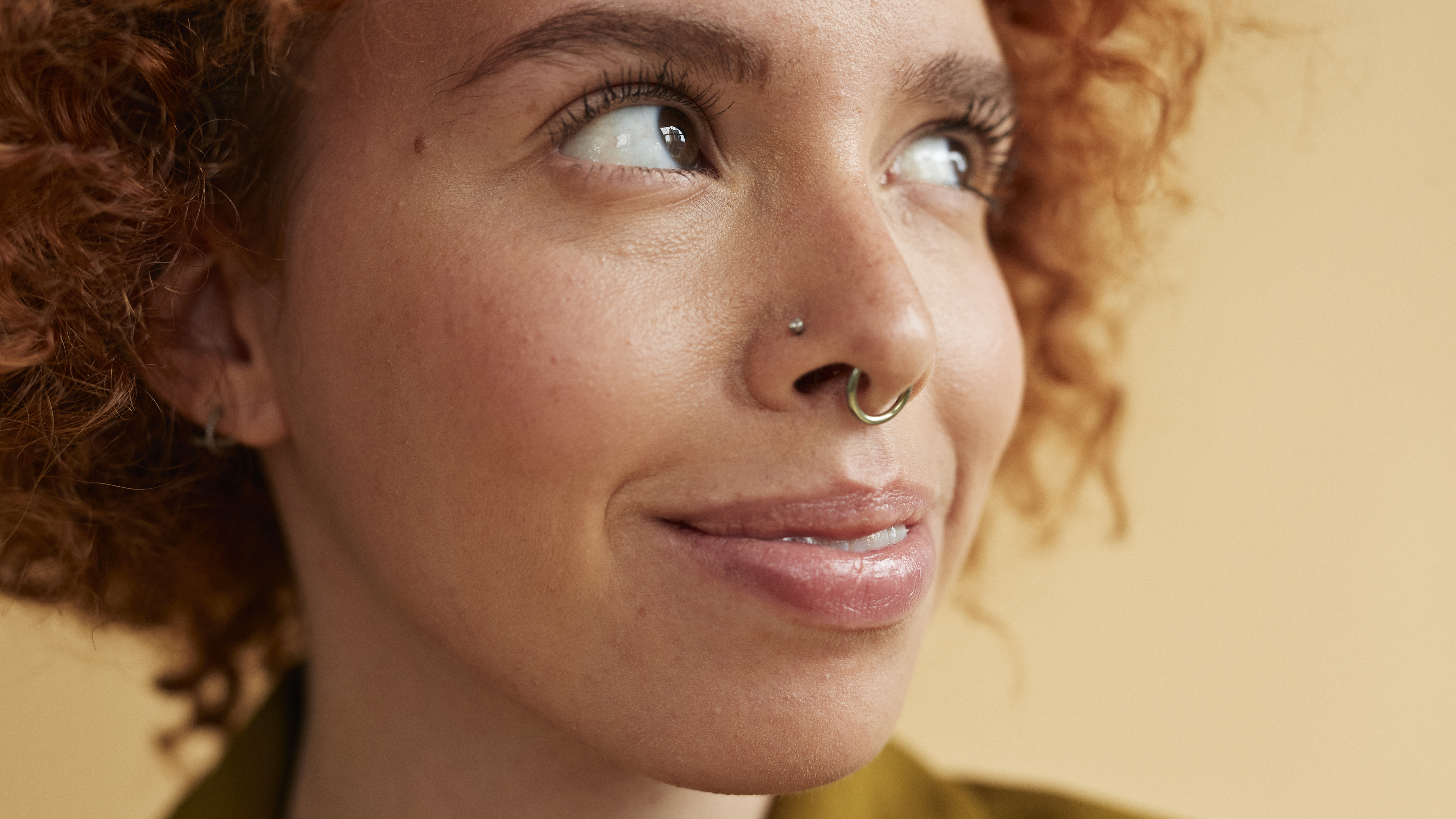 Close-up of smiling young redhead non-binary person with autism and AuDHD with nose piercing looking up against beige background
