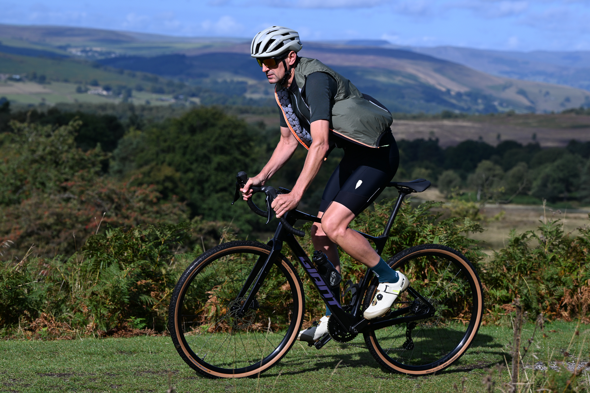 Man wearing black shorts, green jersey and a green gilet riding a black gravel bike pas the camera, right to left