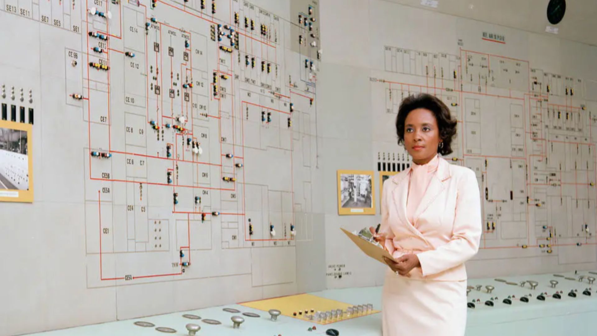 A Black woman wearing a pale pink skirt suit set stands in front of a control board with diagrams.