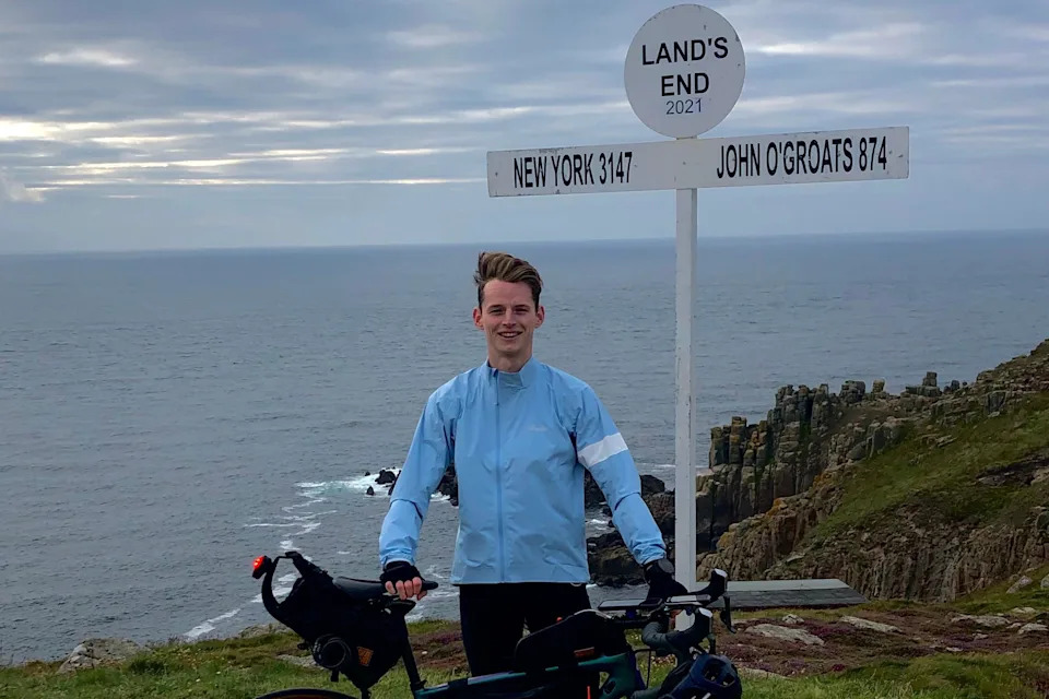 Tom Williamson in a blue jacket holding up a bicycle and smiling for the camera. He is next to a sign that reads Land's End and points towards New York with a distance of 3147 listed and towards John O'Groats with 874 listed. The sea is visible behind him.