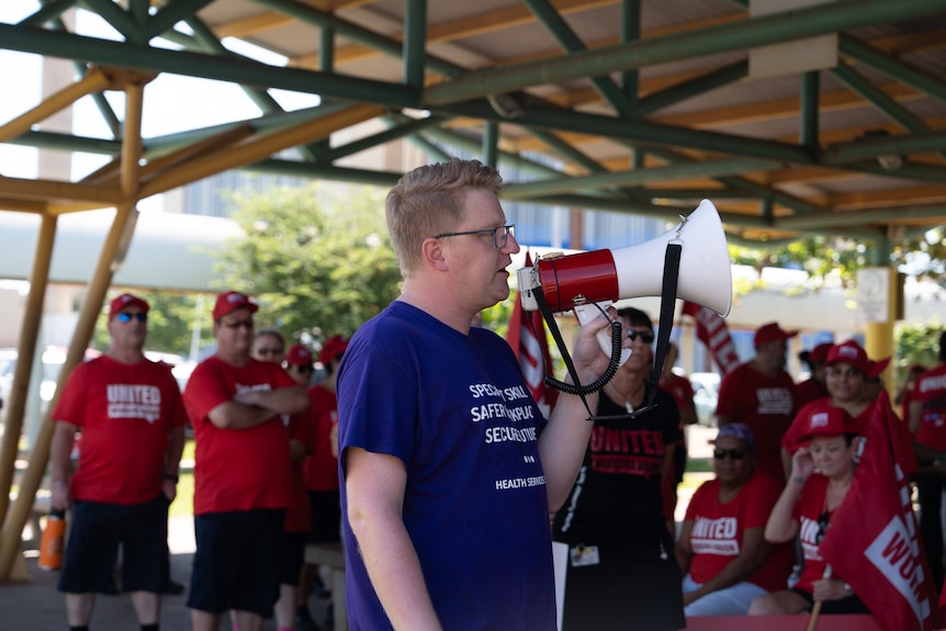 A white man in blue shirt standing in centre, holding a white / red megaphone, crowd of people in red shirts behind him.