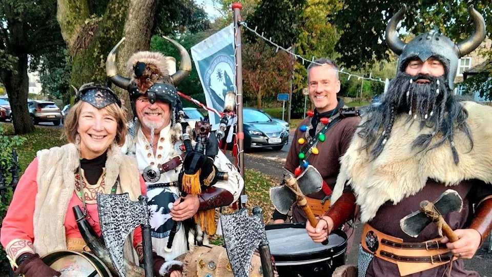 Four of the Biking Vikings stand in a line in a grassy area and smile brightly at the camera. They are wearing various Viking outfits including helmets with horns, furs and accessories with patterns on them. They are also holding their instruments. Including the pipes and drums. One Viking at the front is holding two small axes.
