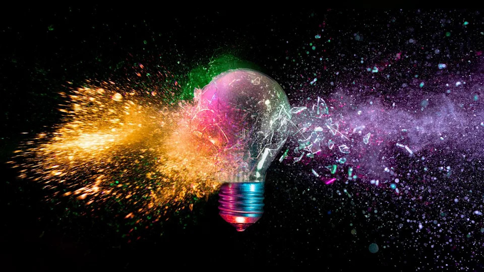 A lightbulb is shattered with various colors coming out of it against a black background.