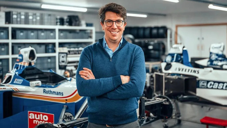 Jonathan Kennard has short brown hair and glasses, and is wearing a blue quarter-zip with his arms crossed. Behind him is a workshop with old F1 cars in.