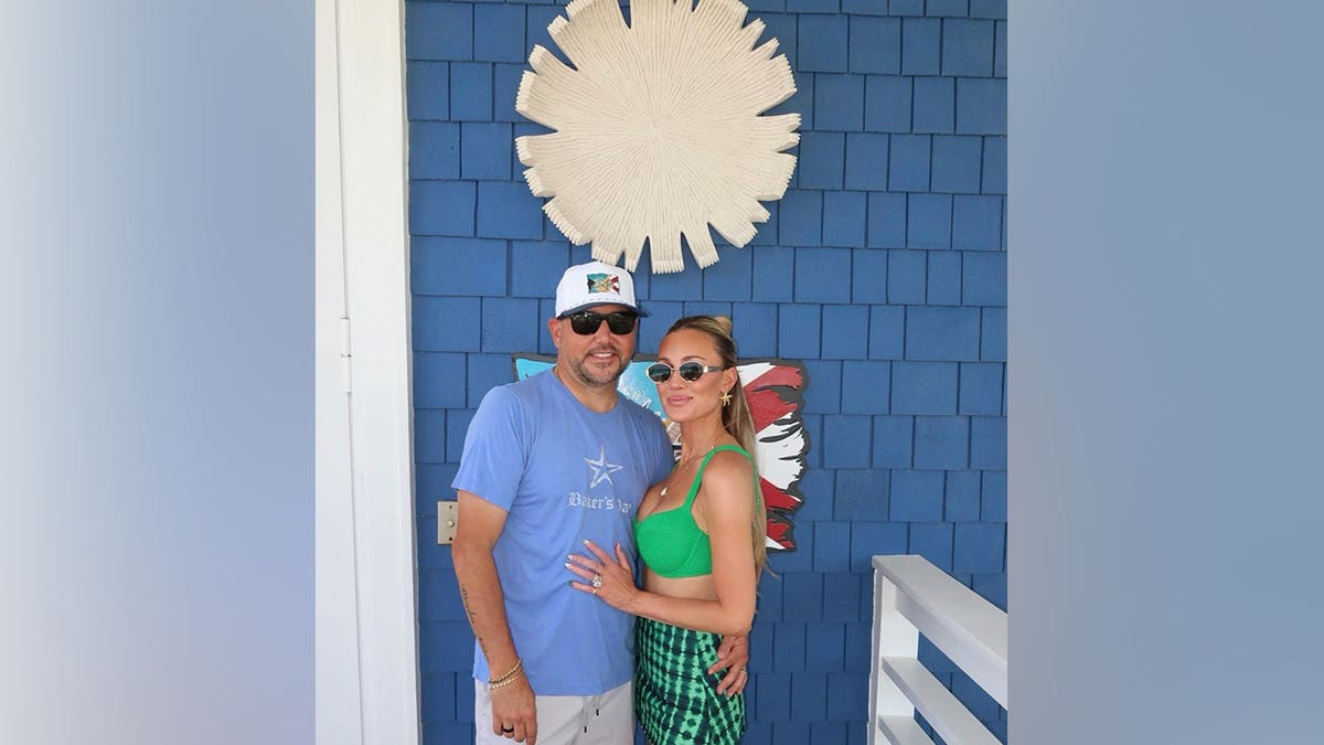 Brittany Aldean poses beside husband Jason Aldean while wearing a green bikini top and matching patterned skirt in front of a bright blue exterior wall.