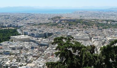 Panoramic view of Athens from above, with the Acropolis visible in the center and dense urban housing stretching toward the sea.