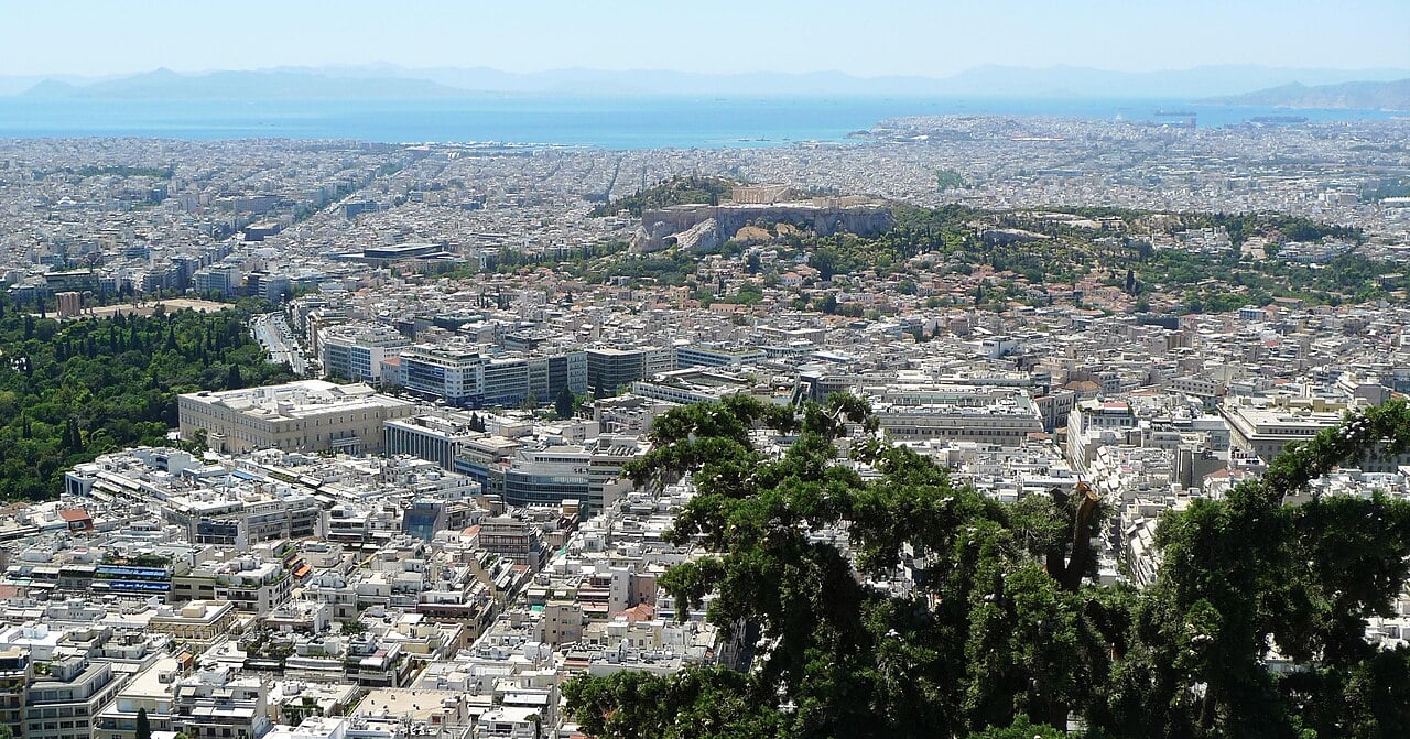 Panoramic view of Athens from above, with the Acropolis visible in the center and dense urban housing stretching toward the sea.