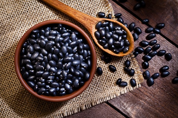 Top view of a brown bowl filled with black beans shot on rustic wood table. A wooden spoon is beside the bowl with some beans on it.