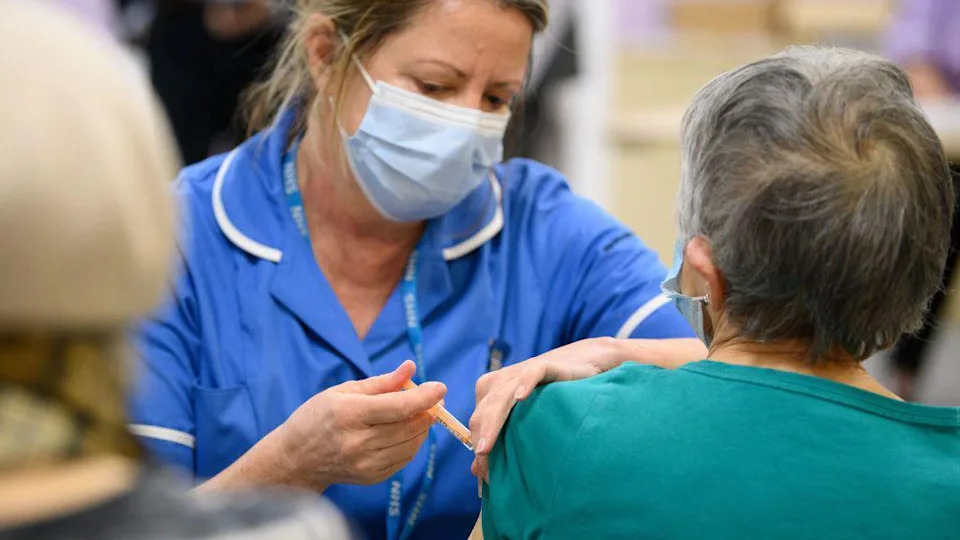 The image shows a healthcare setting where a medical professional is administering an injection to another person’s upper arm. The medical professional is wearing a blue uniform, a face mask, and holding a syringe carefully while preparing or delivering the shot. The seated individual has their sleeve slightly pulled up to expose the upper arm for the injection. The background appears softly blurred, with other people and elements suggesting a busy clinic or vaccination site.