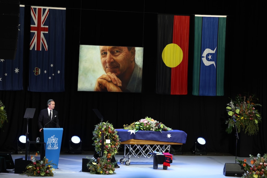 A casket on the stage at the state funeral for Neale Daniher. A portrait of Neale hangs in the background.