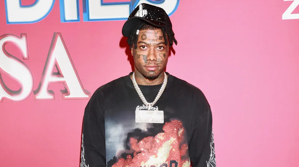 Blueface wearing a black shirt and a chain necklace stands in front of a pink background.