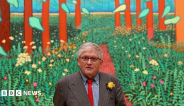 A man with grey hair and wearing black-rimmed glasses is standing in front of a colourful painting depicting green plants and tall trees. He is wearing a grey pin-striped suit, blue shirt and red tie with a yellow flower in his lapel.