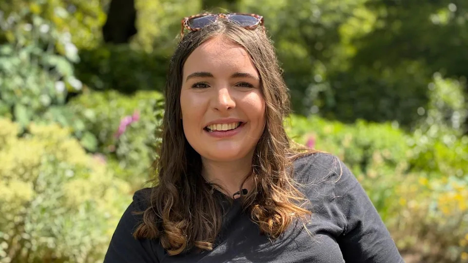 A woman in her late twenties with long brown hair, wearing a black top, smiling at the camera. She's sat in a public park with greenery behind her.