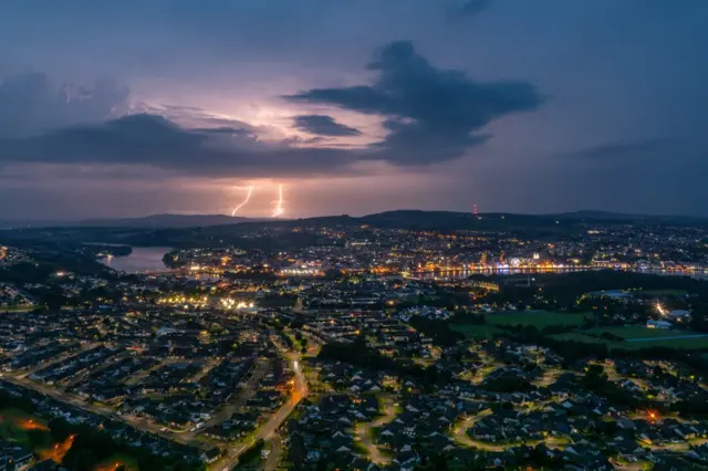 Two lightning strikes can be seen behind a dark cloud in the distance above the county of Londonderry. The city is lit up by street lights in the front of the picture