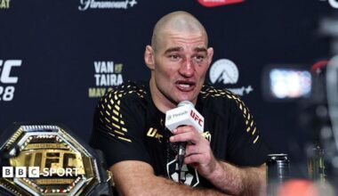 Sean Strickland, wearing a black t-shirt with gold pattern on the shoulders, sits talking into a white and black microphone, held in his left hand, with a gold octagonal UFC belt to his right