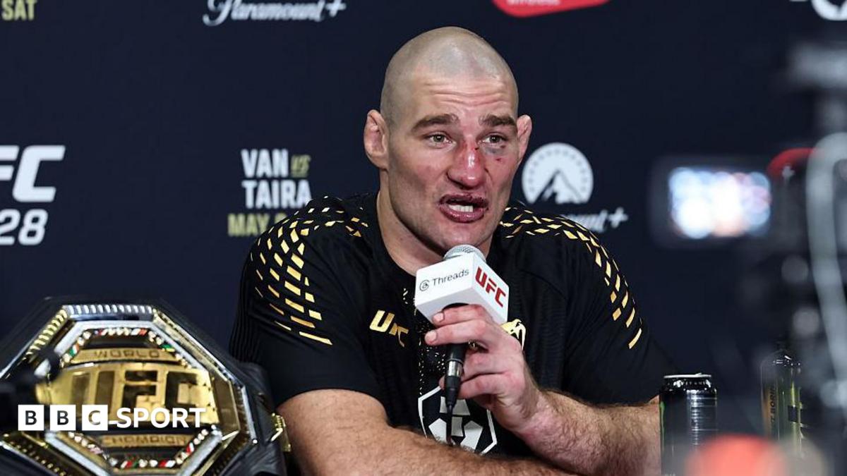 Sean Strickland, wearing a black t-shirt with gold pattern on the shoulders, sits talking into a white and black microphone, held in his left hand, with a gold octagonal UFC belt to his right