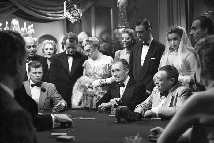 Black-and-white still of Barry Nelson as James “Jimmy” Bond at a casino table, surrounded by tuxedoed gamblers and elegantly dressed women during a tense card game in 1954’s Casino Royale.