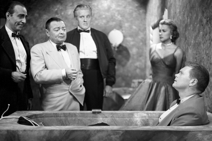 Peter Lorre as Le Chiffre stands in a white dinner jacket beside Barry Nelson’s James Bond, who is slumped in a bath, as Linda Christian’s Valerie Mathis watches anxiously from the wall behind.