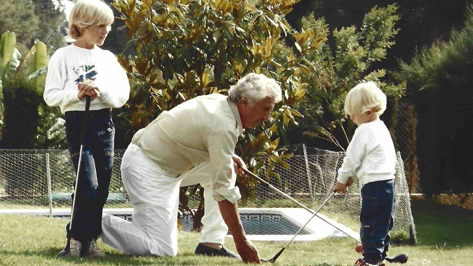 An older man kneels on the grass, helping a young child grasp a golf club—his secret weapon for fun—while another child stands nearby holding a club. They are outside near a pool, surrounded by lush greenery.