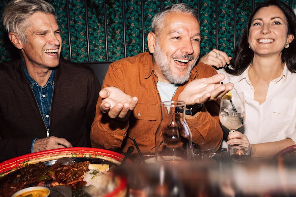 Three people share a joyful moment at a dining table, laughing and conversing, with food and drinks in front of them