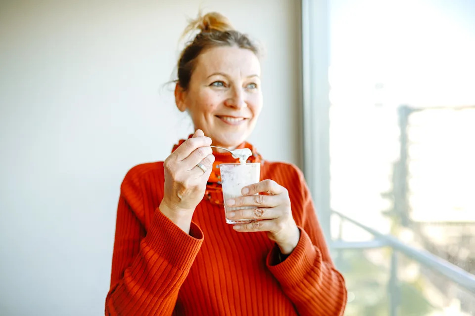 Person smiling, holding a yogurt and spoon near a window. They are wearing a ribbed sweater and have their hair tied up