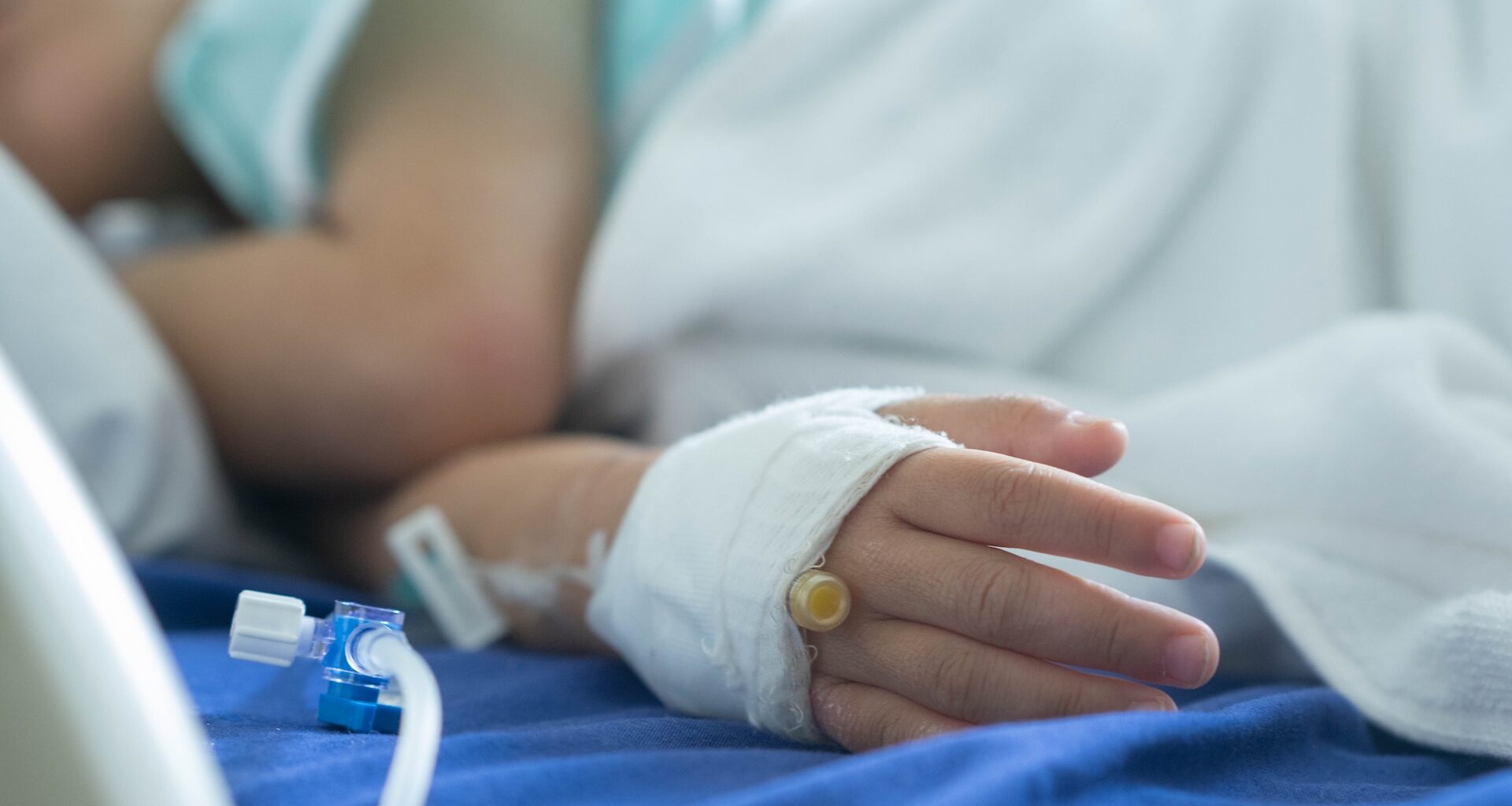 A close-up of a child's hand with a bandage and IV line in a hospital setting.