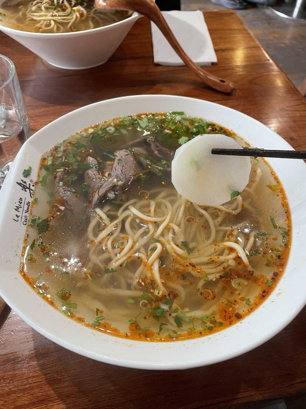 Close-up of beef noodle soup at Le Mein Craft Noodle, showing noodles, sliced beef, and fresh herbs, with chopsticks holding a piece of radish above the bowl.
