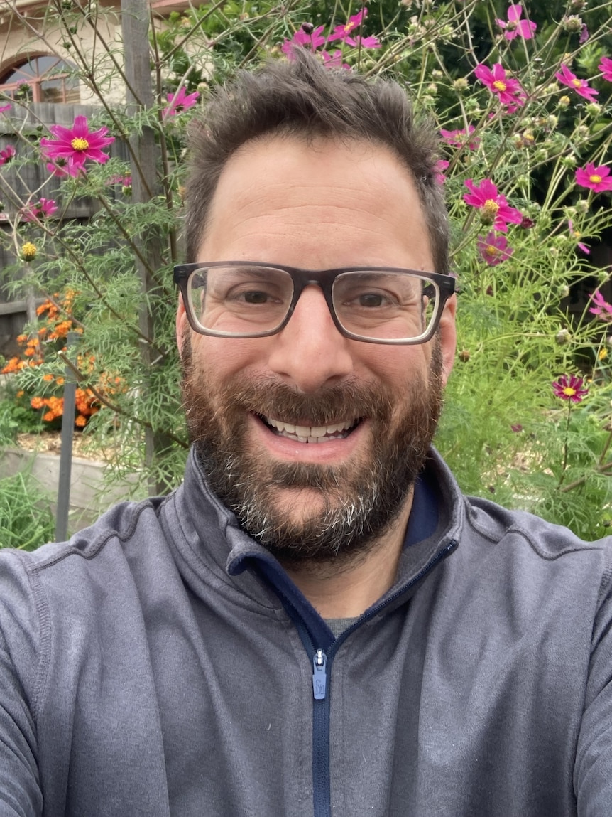 A smiling man with standing in a community garden, with pink cosmos flowers in the background.