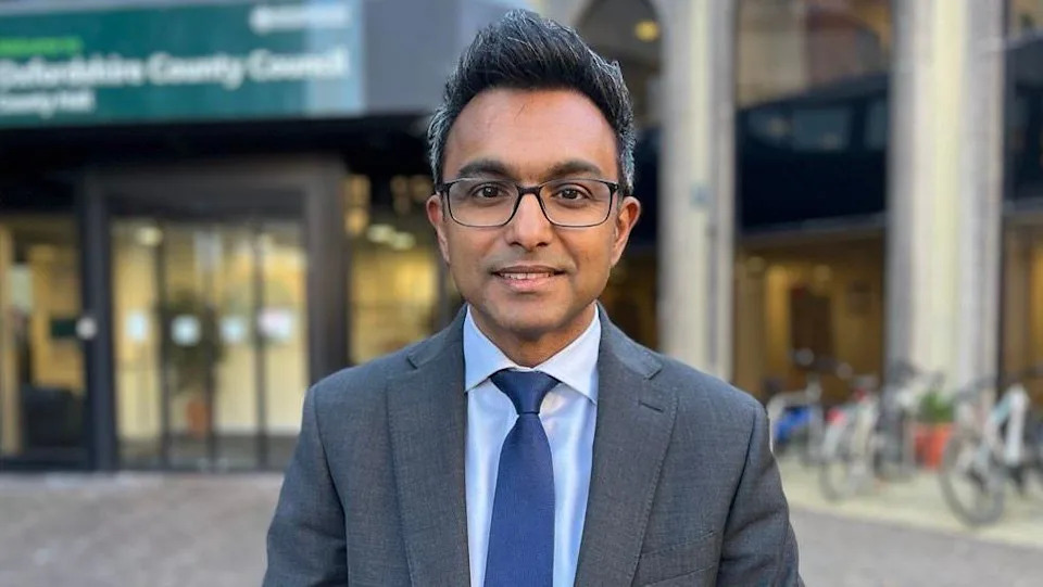 Ansaf Azhar outside the council headquarters. He has short greying black hair, wears glasses, a grey suit, a light blue shirt and a dark blue tie.