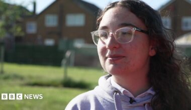 A picture of Mary, she has dark brown curly hair and is wearing glasses. There are houses in the background.