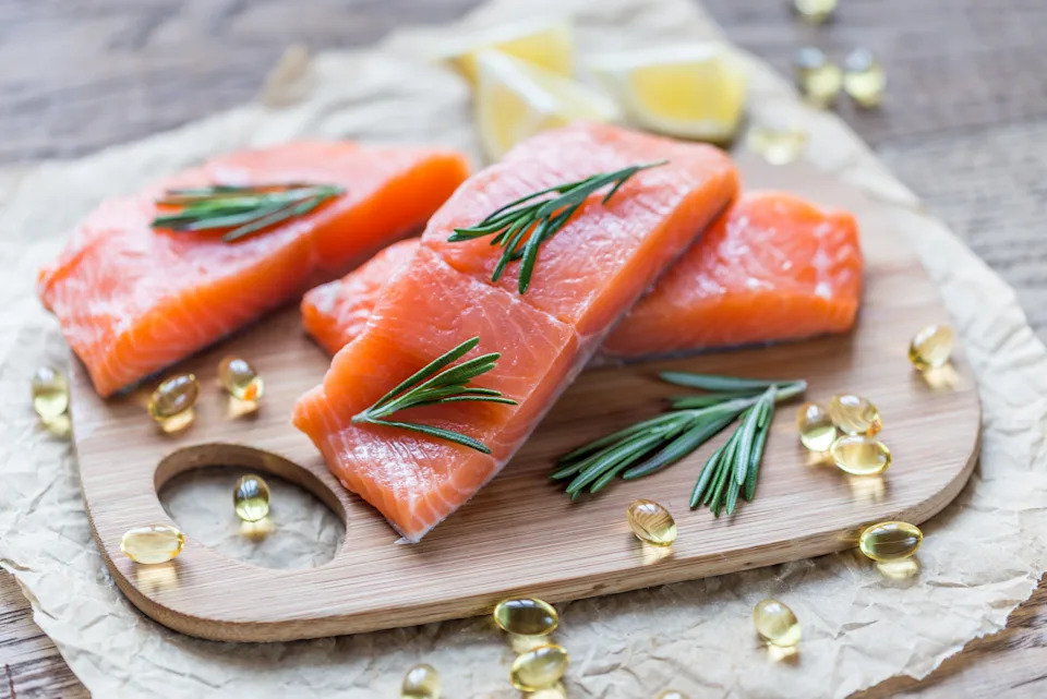 Salmon fillets with rosemary on a cutting board, surrounded by fish oil capsules and lemon wedges