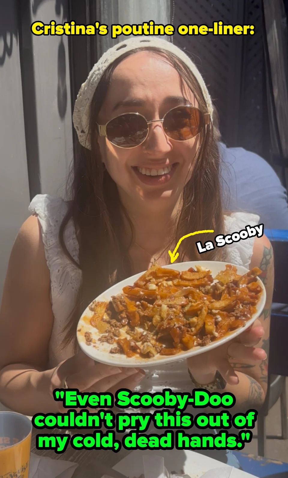 Person with short hair sitting outdoors, holding a plate of food, smiling gladly at it. People are seated at tables in the background