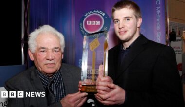 Two people standing indoors at an event booth display branded ‘BBC Midlands’ in the background. Both individuals are holding a tall, transparent trophy featuring a golden microphone design mounted on a base with engraved text. The backdrop includes a purple gradient panel with a circular BBC Sports logo and vertical ‘BBC Midlands’ text along the side. The setting appears to be an awards or media event, with exhibition stands and signage visible in the background.