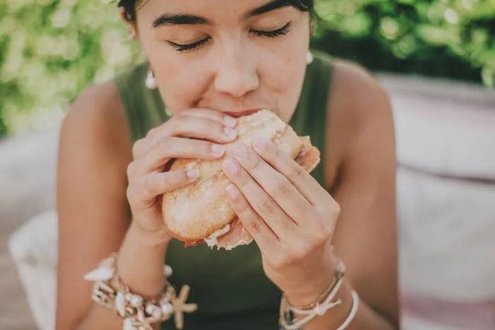 Person enjoying a sandwich with eyes closed, wearing bracelets and sitting outdoors
