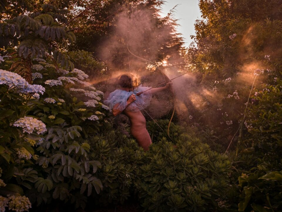 a photograph by Tamara Dean of an individual in a bush taking aim with a cross bow, while wearing a frilly blue tutu around their neck
