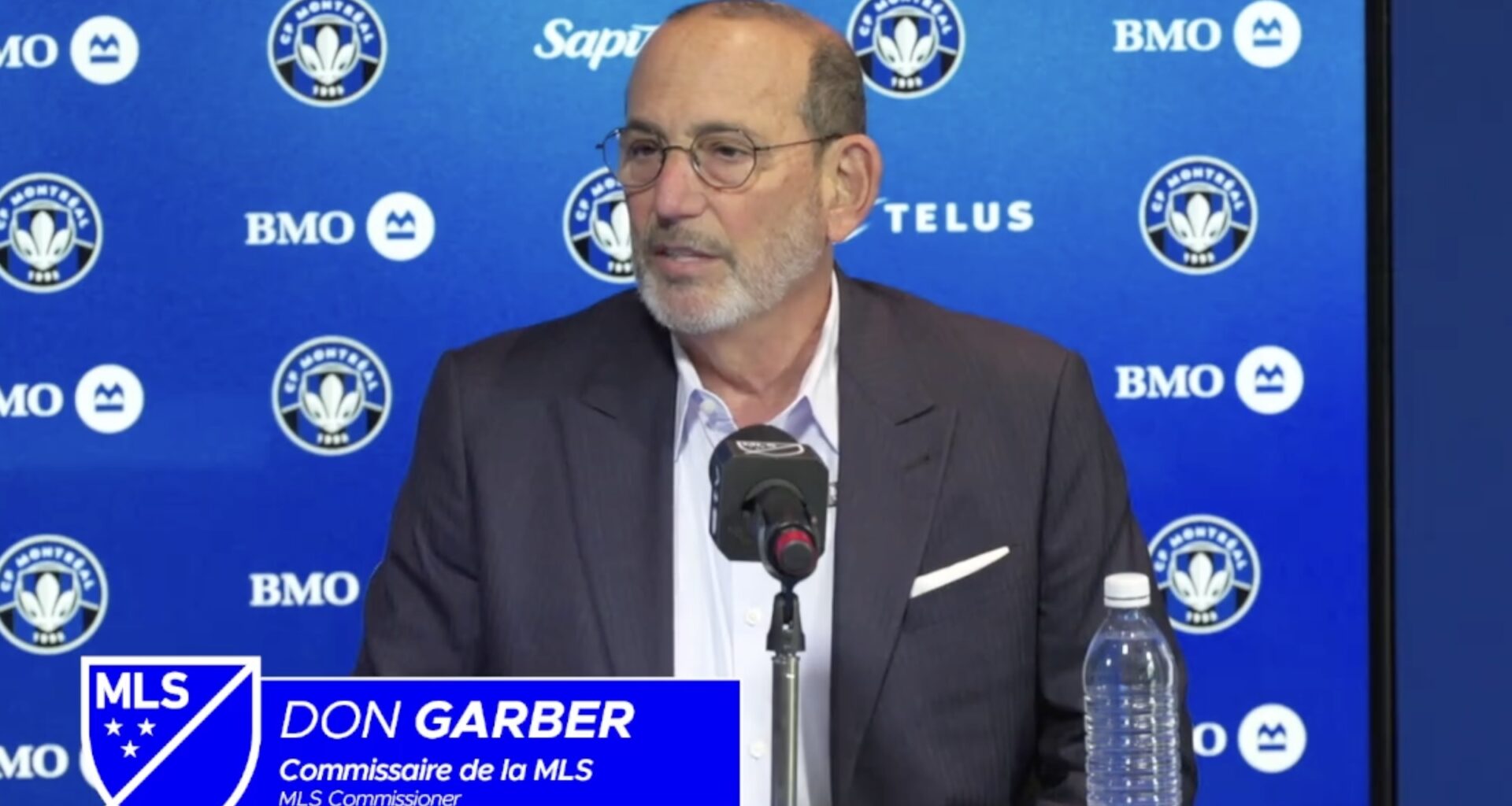 Don Garber speaks while sitting at a desk with a microphone. A graphic superimposed on the image with the MLS logo identifies him. A CF Montréal backdrop is behind.