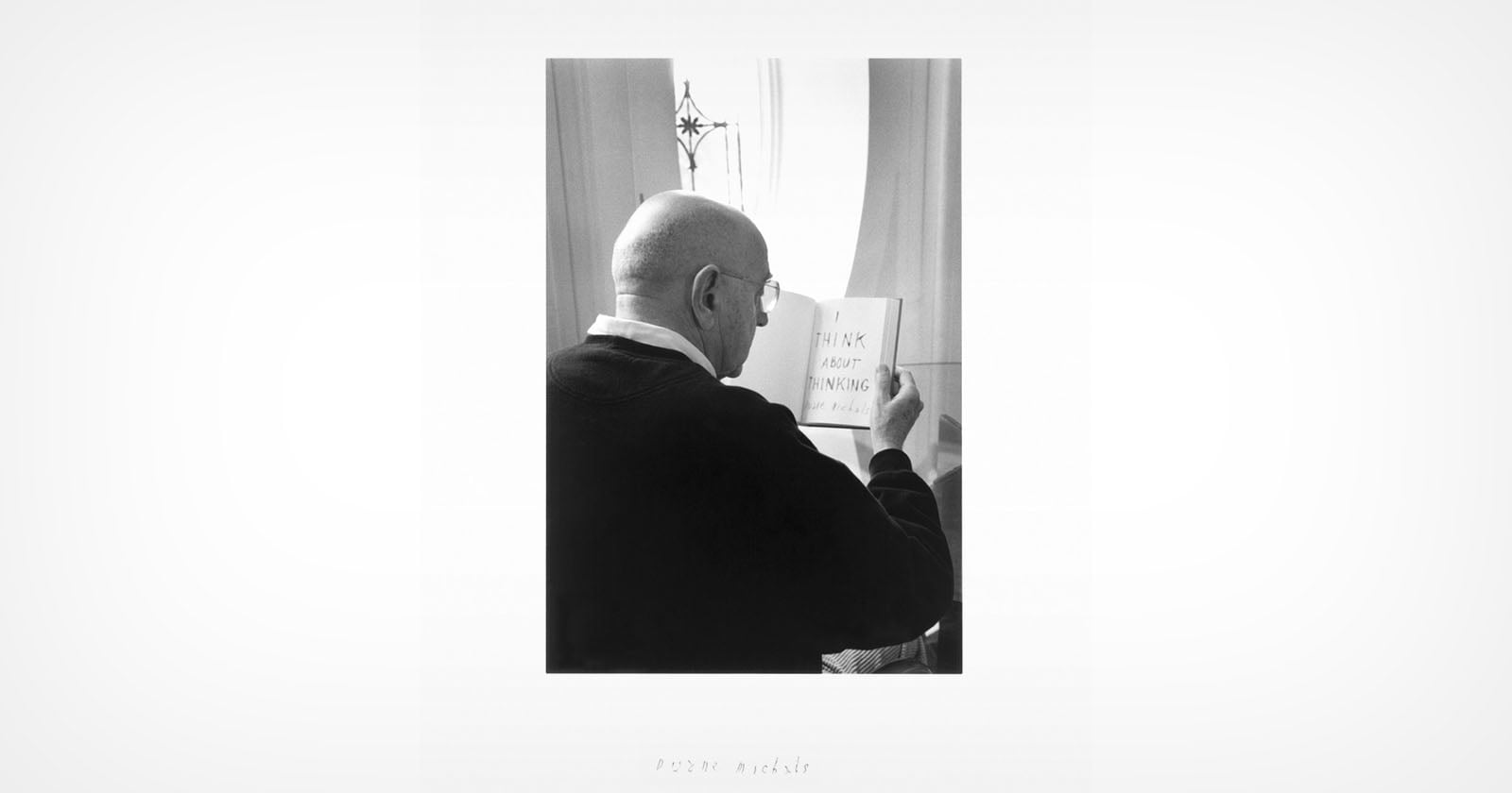 A bald person wearing glasses reads a small book with the words "THINK ABOUT SOMETHING" written on the open page, standing near a window with light streaming in.