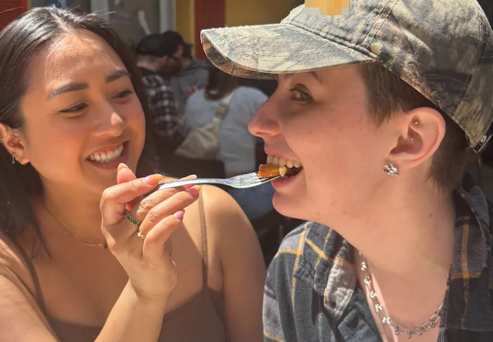 Person smiling feeds another bite from a fork outdoors; both wear casual tops and one has a camouflage hat