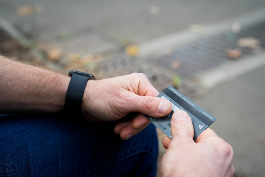 A man wearing jeans holds a grey credit card between his fingers as he sits on a bench.